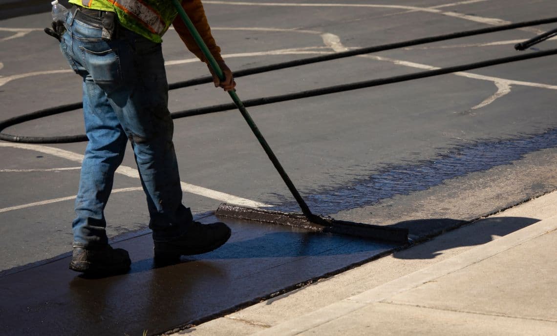 A construction worker in jeans and a safety vest uses a squeegee to apply black asphalt sealcoat evenly to a parking lot surface. The fresh, dark sealant contrasts with the lighter, unsealed asphalt and concrete sidewalk. This image shows the process of protective pavement maintenance.